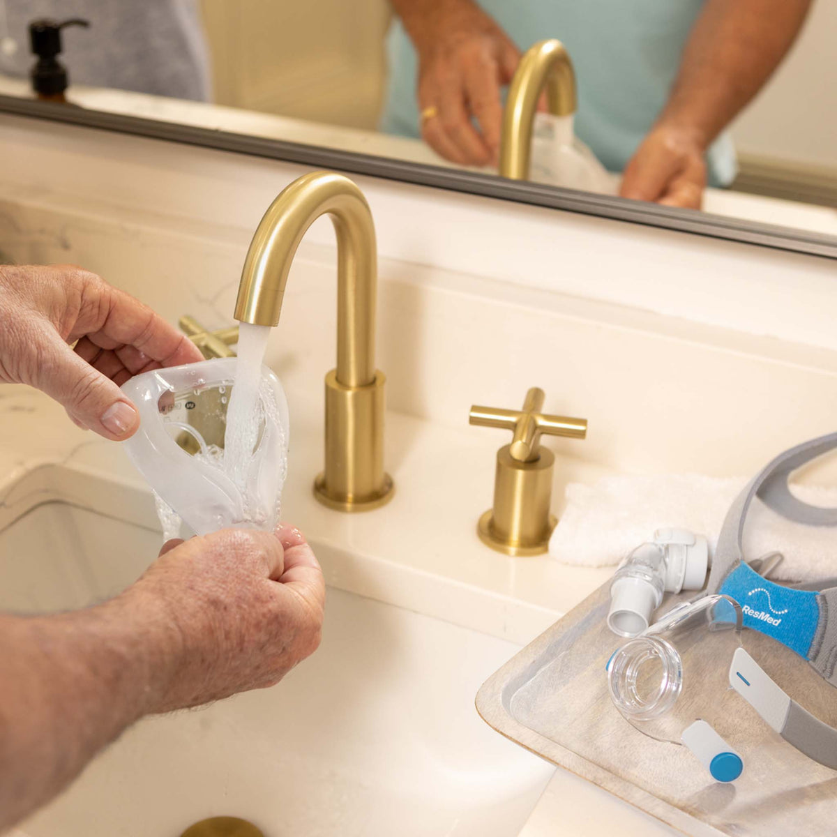 Person washing a clear ResMed CPAP mask cushion under a gold bathroom faucet.