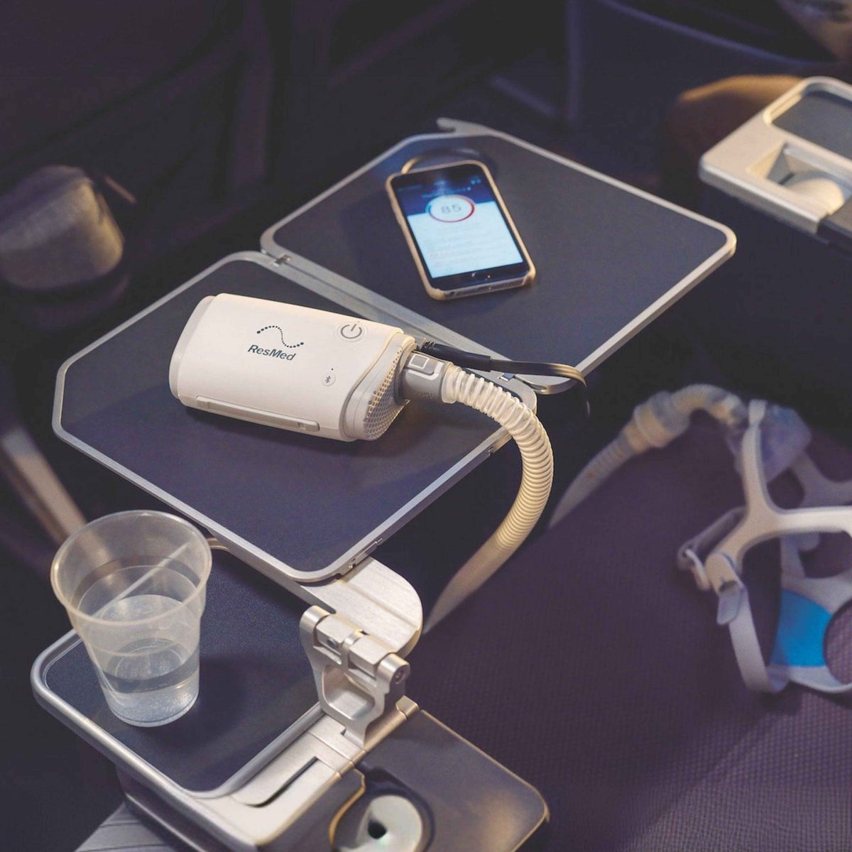 ResMed AirMini travel CPAP machine on an airplane tray table with a smartphone and water cup.