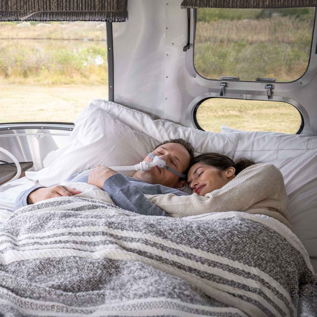 A man using a ResMed CPAP mask while sleeping next to a partner inside a travel trailer.