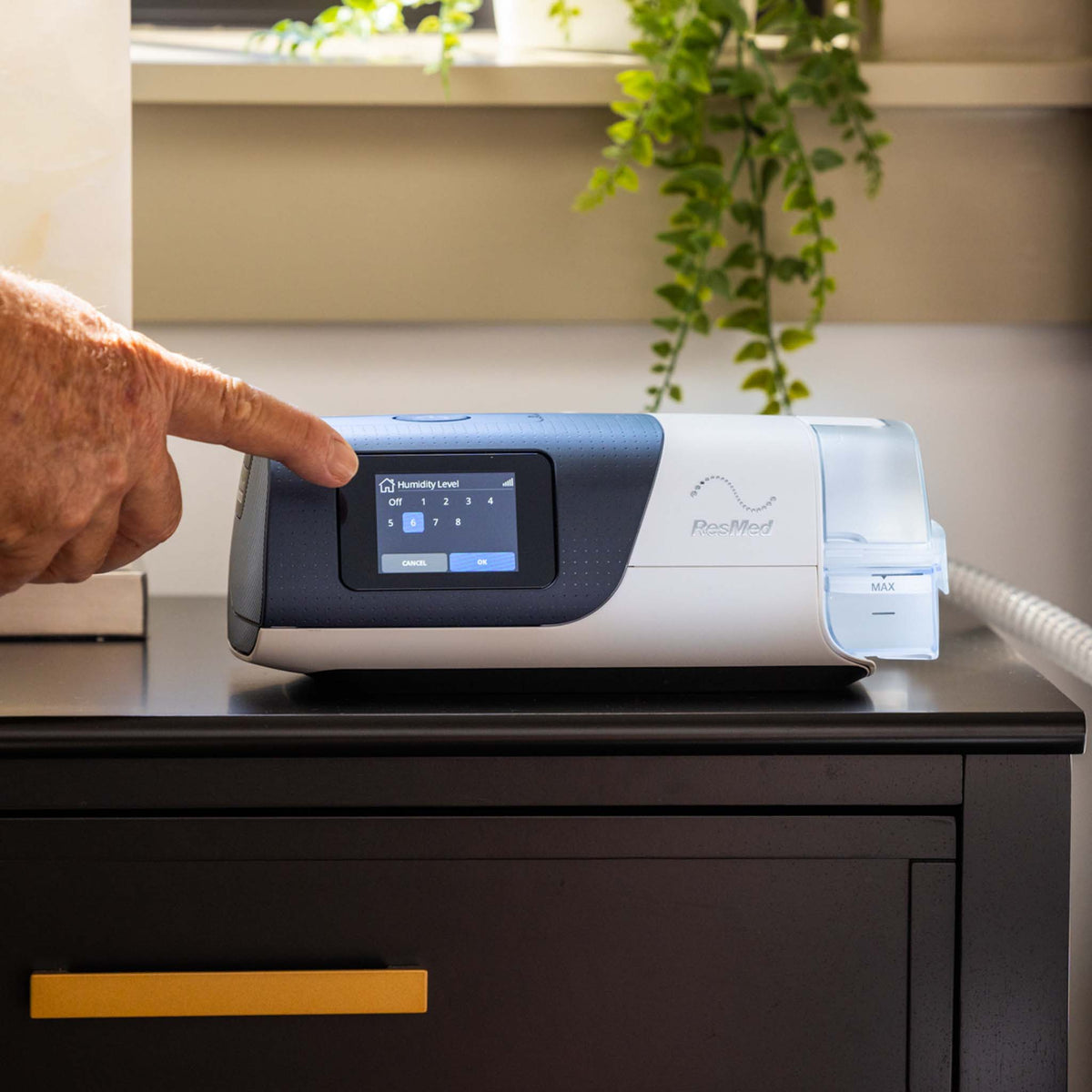 A person adjusting the humidity level on the touchscreen display of a ResMed AirSense 11 AutoSet CPAP machine, which is placed on a dark nightstand with a connected hose.