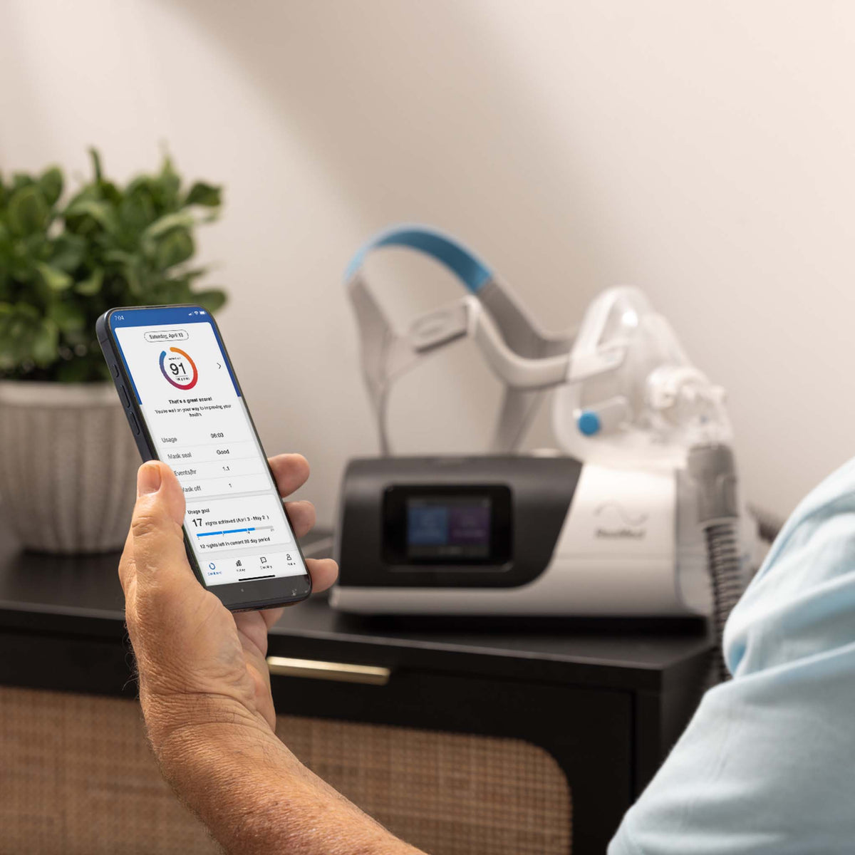 Person holding a smartphone showing sleep data next to a ResMed AirSense 11 CPAP machine.