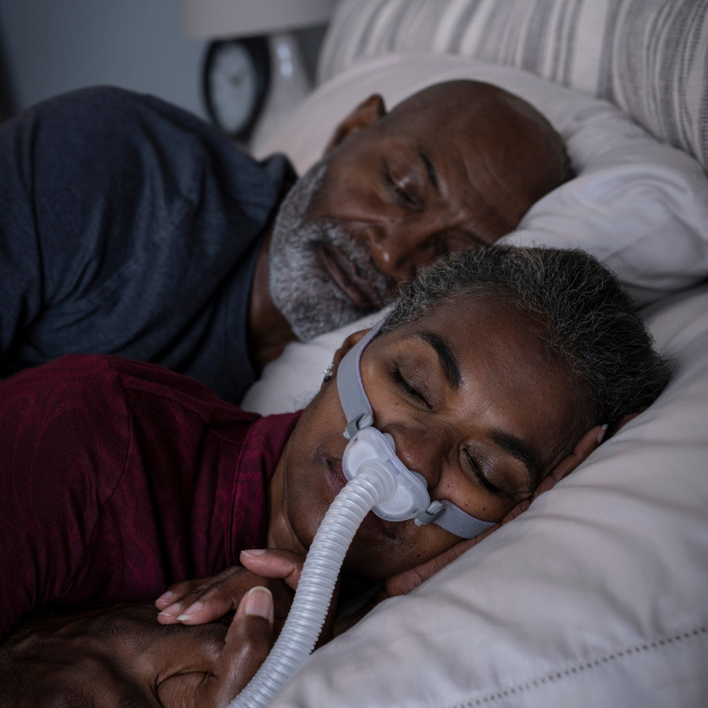 Woman sleeping with a CPAP nasal mask and tubing, lying next to a man in bed.